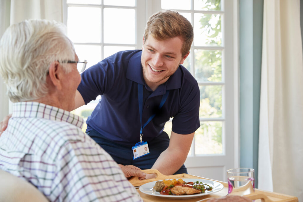 male care worker serving dinner to a senior man at his home